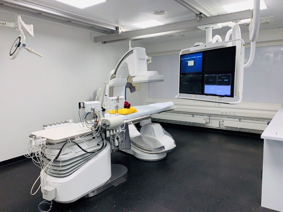 Medizinischer Bildgebungsraum mit Röntgengerät: Patiententisch, grosser Monitor, Deckenarme. Weiss und grau.