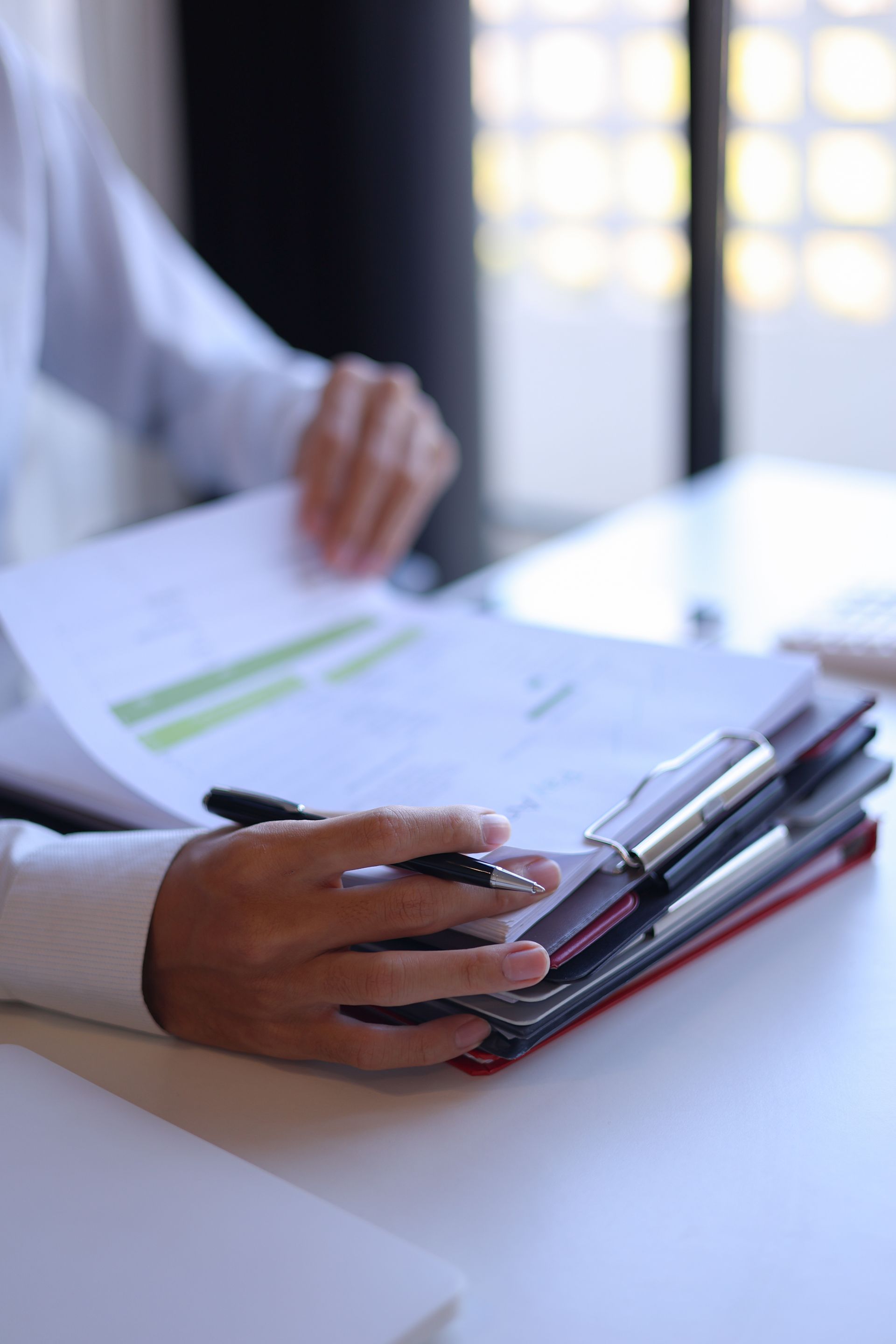 Une personne, assise à un bureau, examine des documents à la main, un stylo à la main. Des feuilles sont posées sur un porte-documents.