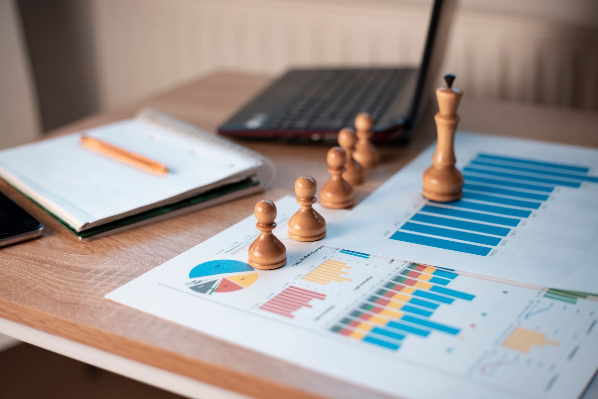 Des pièces d'échecs sur un bureau avec des graphiques, un ordinateur portable, un bloc-notes et un téléphone.