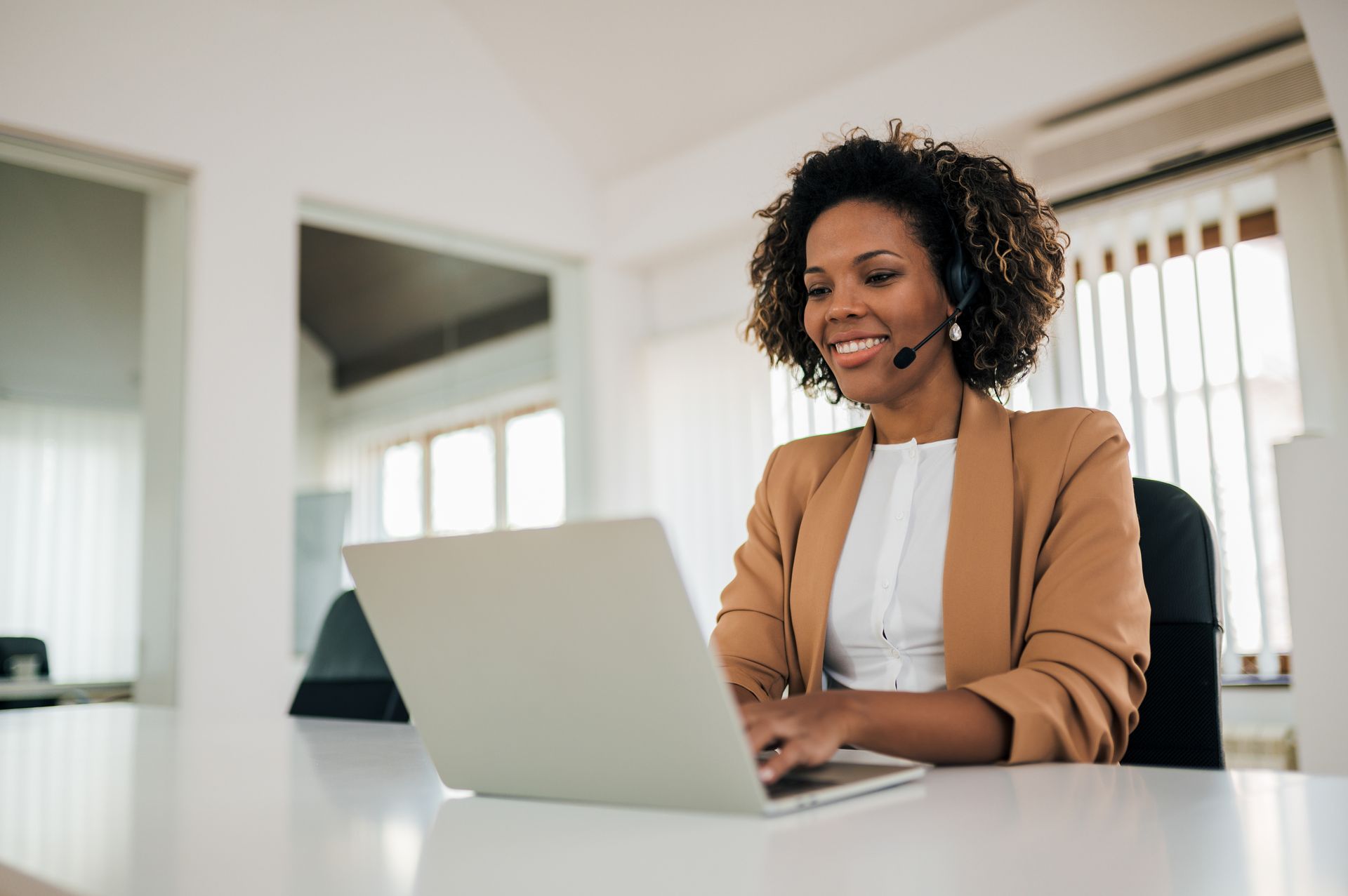 Une femme portant un casque audio sourit en tapant sur un ordinateur portable à un bureau blanc dans un bureau.