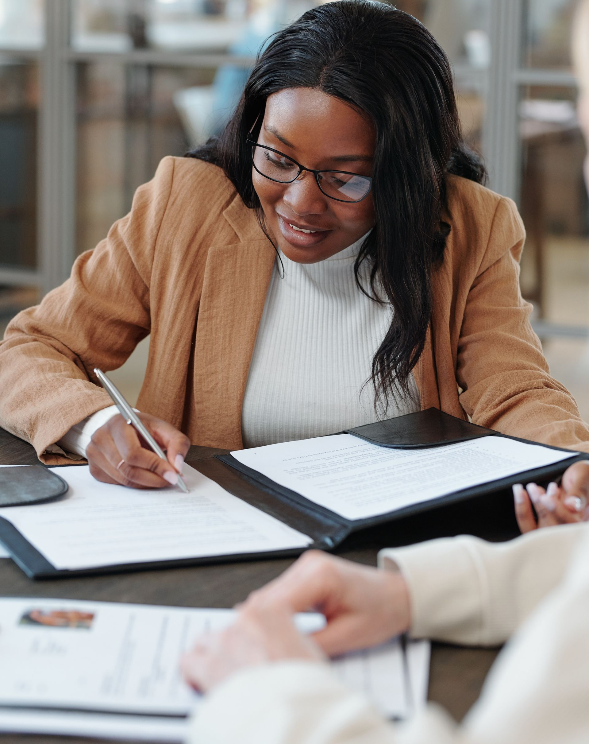 Une femme portant des lunettes et un blazer beige signe des documents à une table.