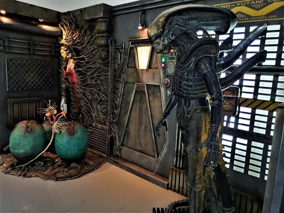 Una estatua de un extraterrestre está parada en una habitación al lado de dos huevos extraterrestres.