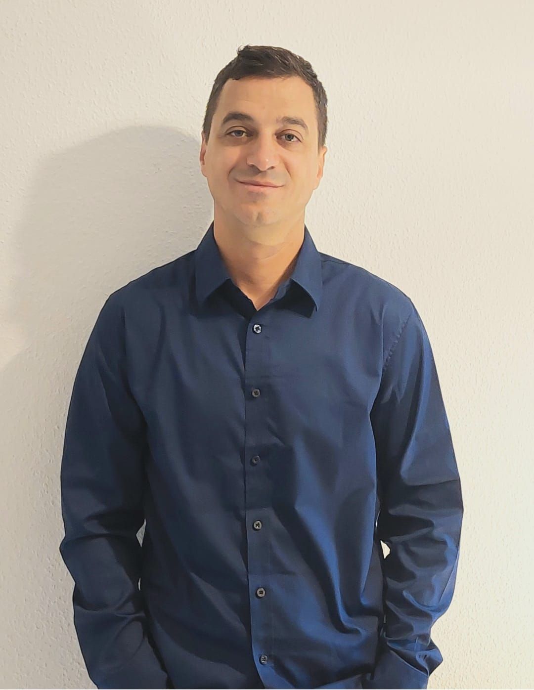 Ein Mann in einem blauen Hemd steht mit den Händen in den Taschen vor einer weißen Wand.