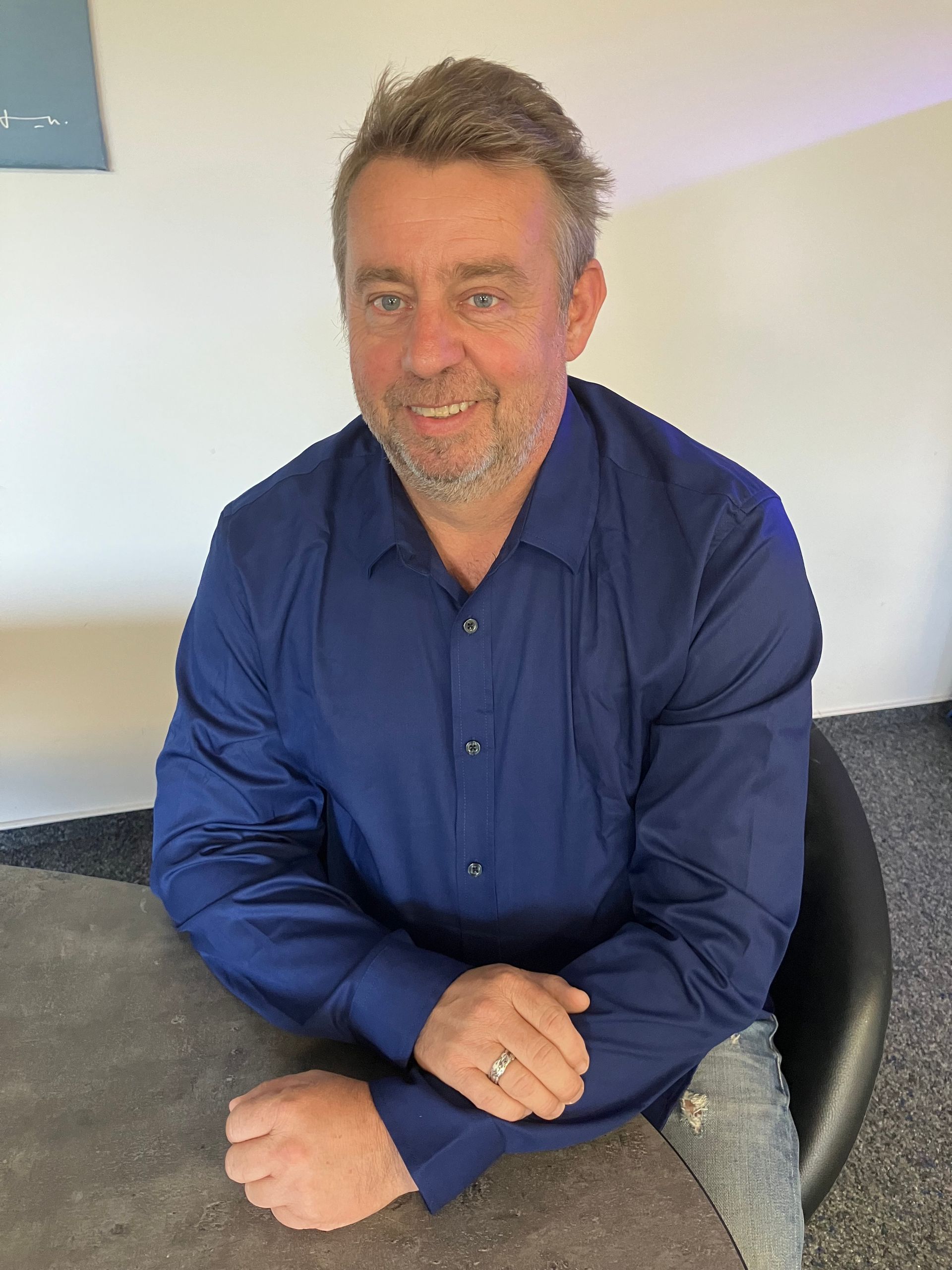 Ein Mann in einem blauen Hemd sitzt mit gefalteten Händen an einem Tisch.