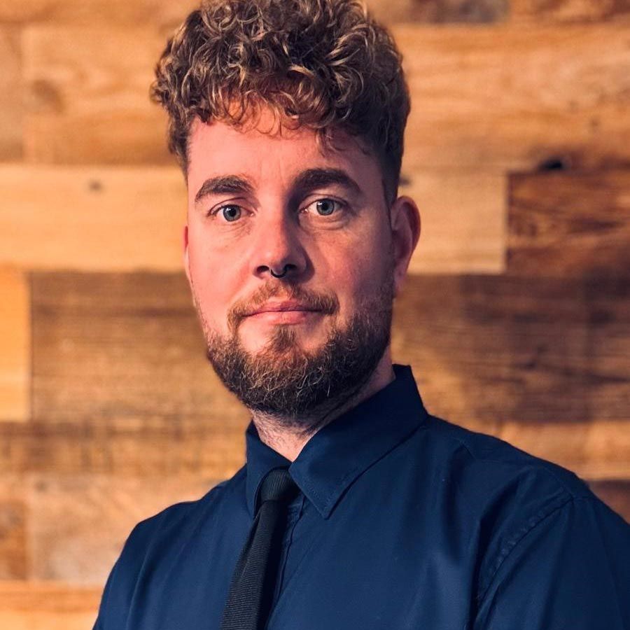 Ein Mann mit lockigem Haar und Bart trägt ein blaues Hemd und eine Krawatte.