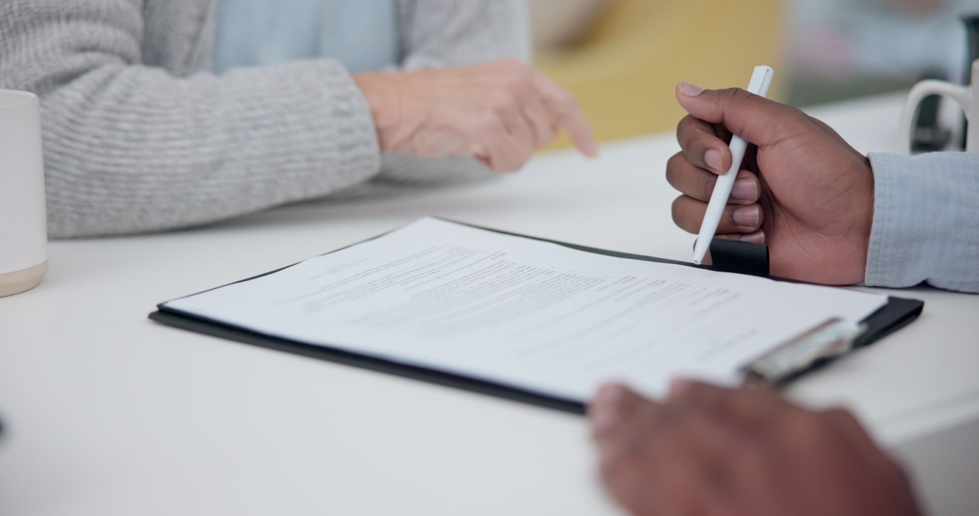 Photo d'un calepin avec feuille blanche dessus, posé sur une table avec deux personnes le signant et le prenant en considération. Une personne tient un stylo. On ne voit que leurs bras.