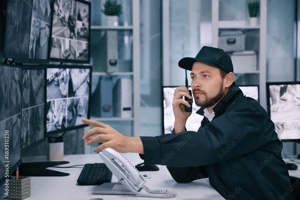 Ein Mann an seinem Büroschreibtisch überwacht die Aufnahmen von Sicherheitskameras auf mehreren Bildschirmen und telefoniert gleichzeitig.