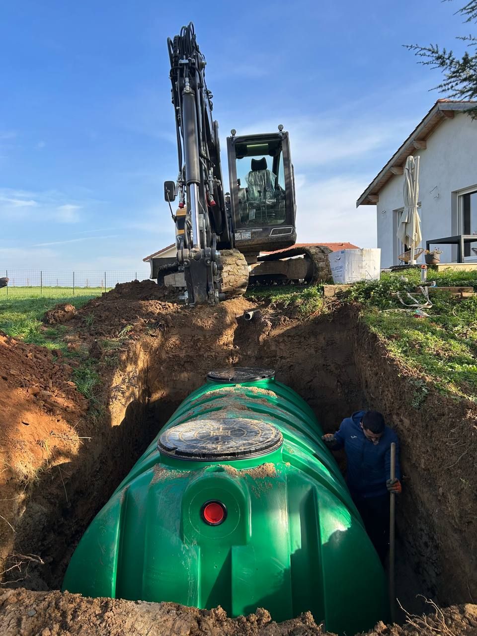 Une excavatrice creuse une tranchée à côté d'une cuve souterraine verte près d'une maison dans une zone rurale