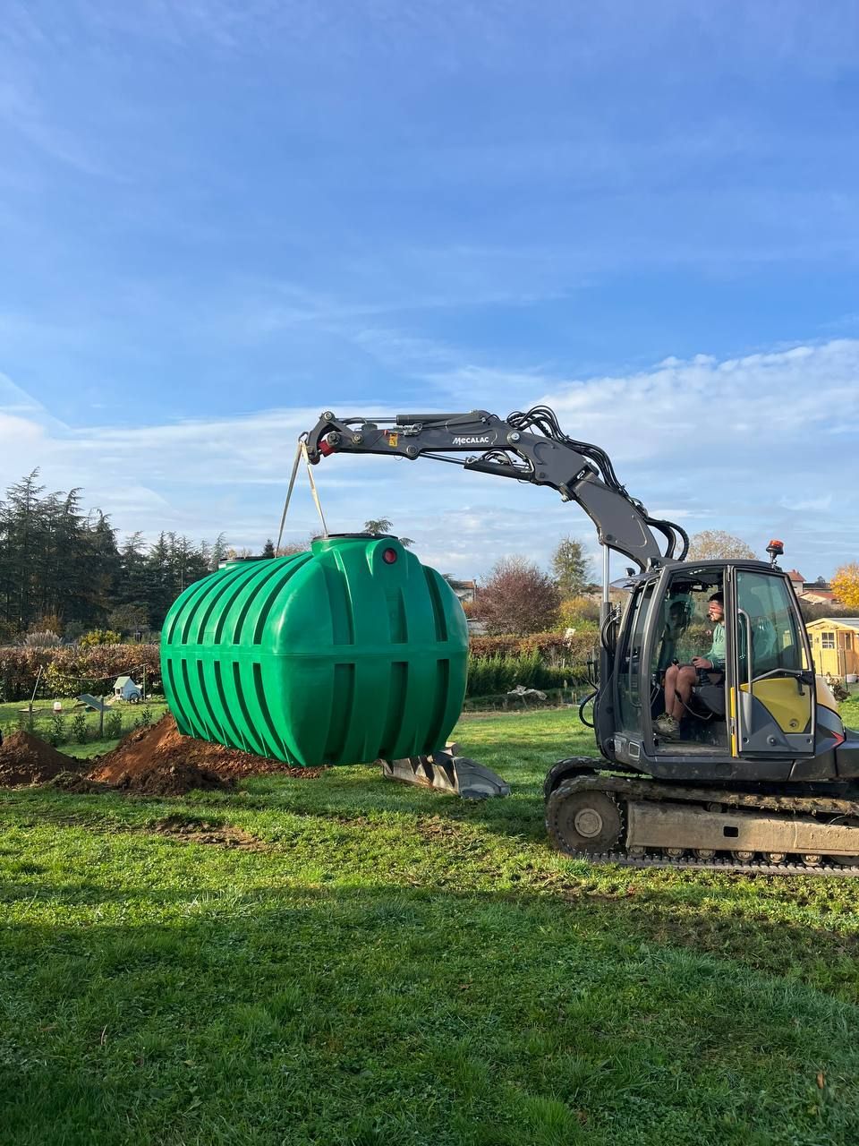 Une excavatrice soulève un grand réservoir cylindrique vert dans un champ herbeux sous un ciel bleu.
