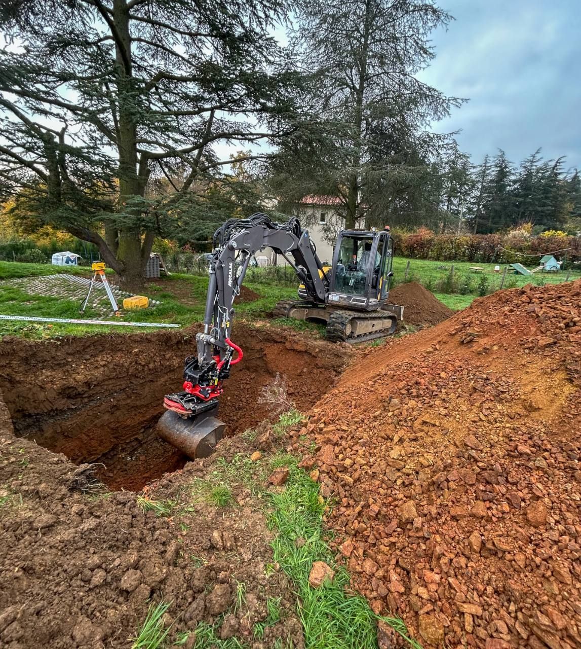 Une pelleteuse creuse une tranchée dans un jardin boueux, à côté d'un grand tas de terre et d'arbres d'automne.