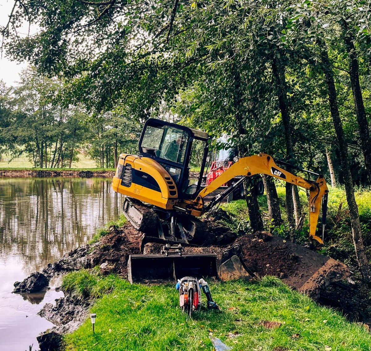 Une pelleteuse jaune creuse la boue au bord d'un étang sous des arbres, avec un conteneur rouge au premier plan