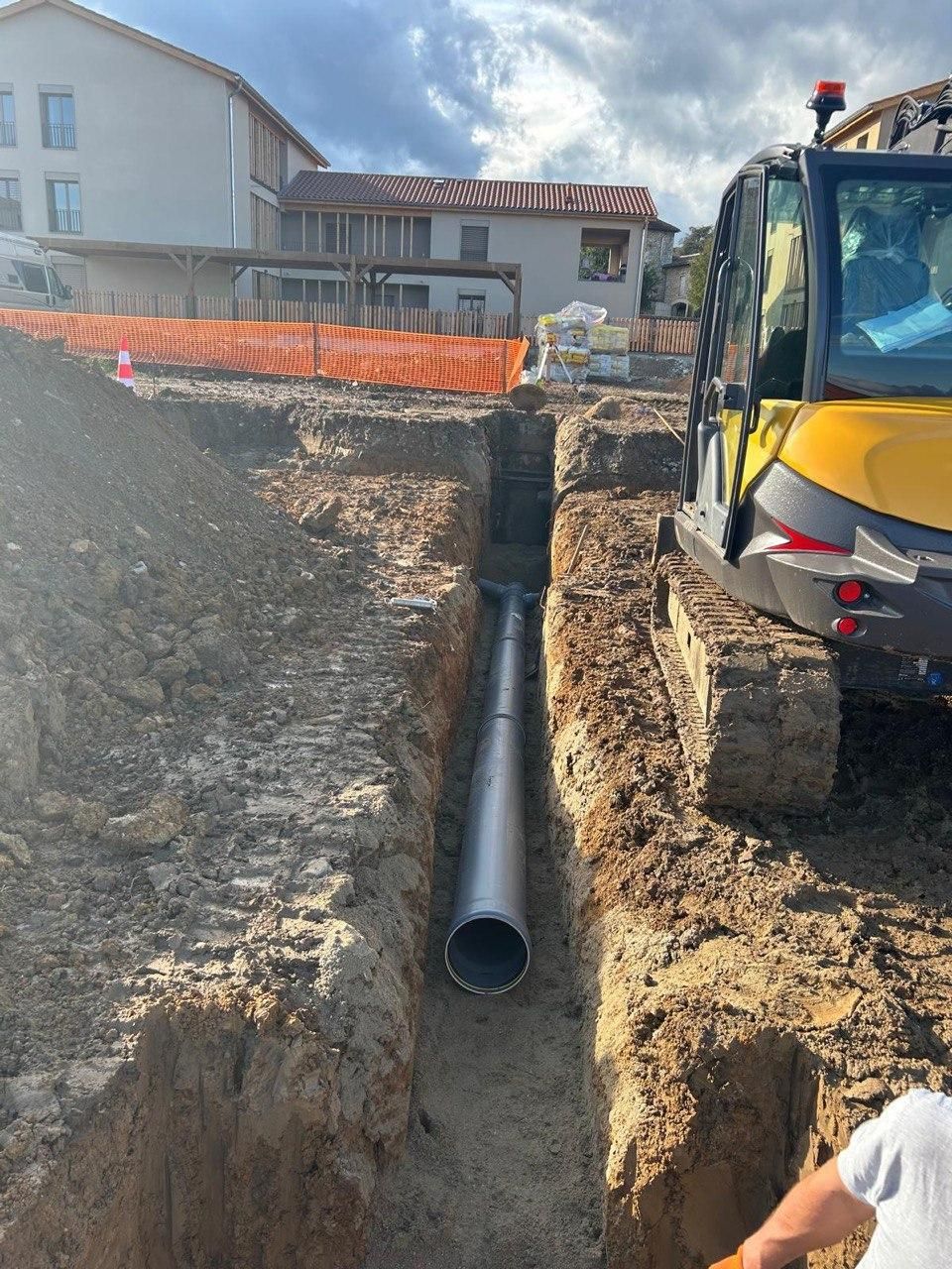 Tranchée de chantier avec un gros tuyau noir à côté d'une excavatrice jaune et une maison en arrière-plan