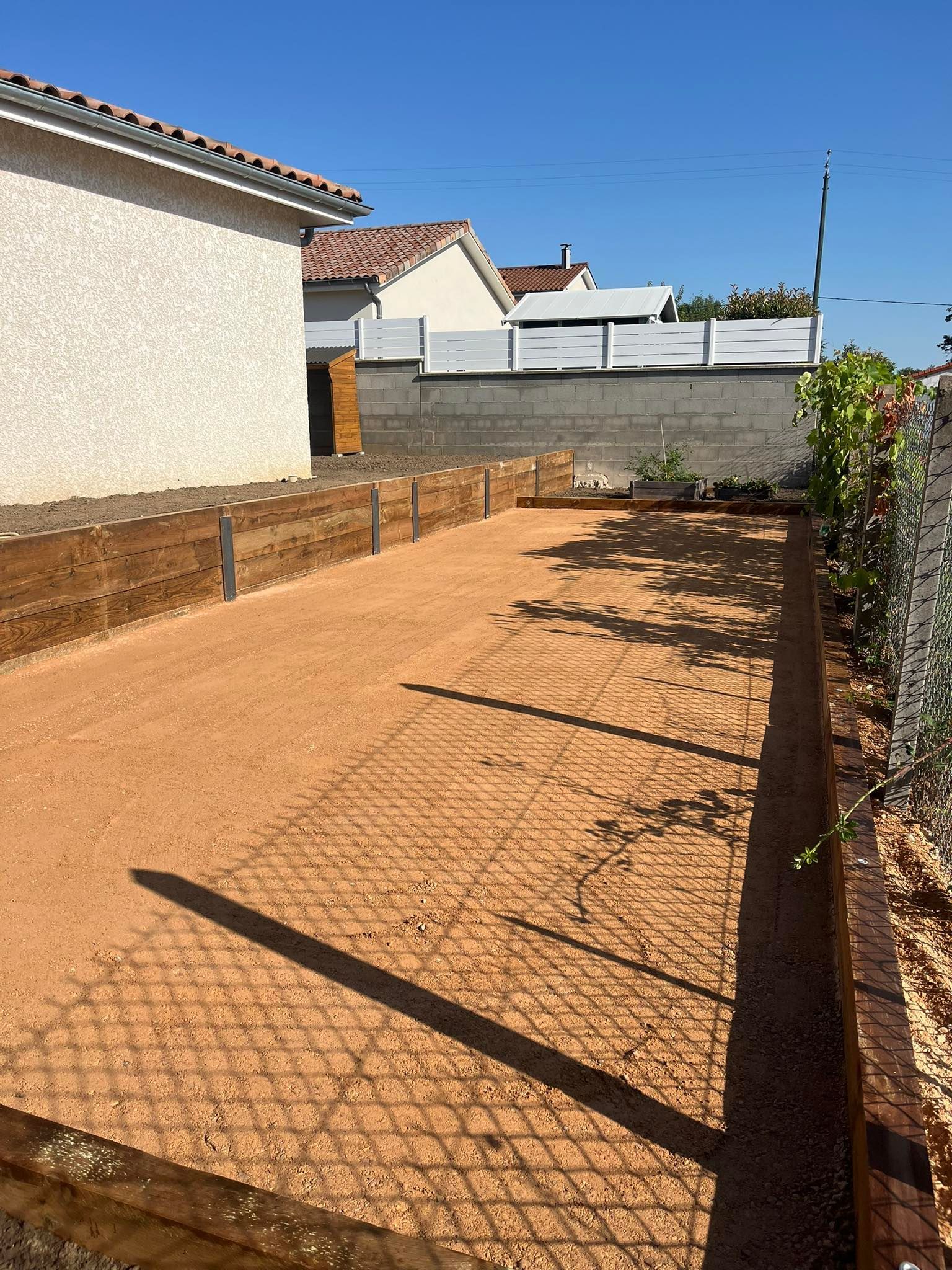 Cour étroite en gravier jouxtant une maison, avec une clôture et des maisons voisines baignées de soleil