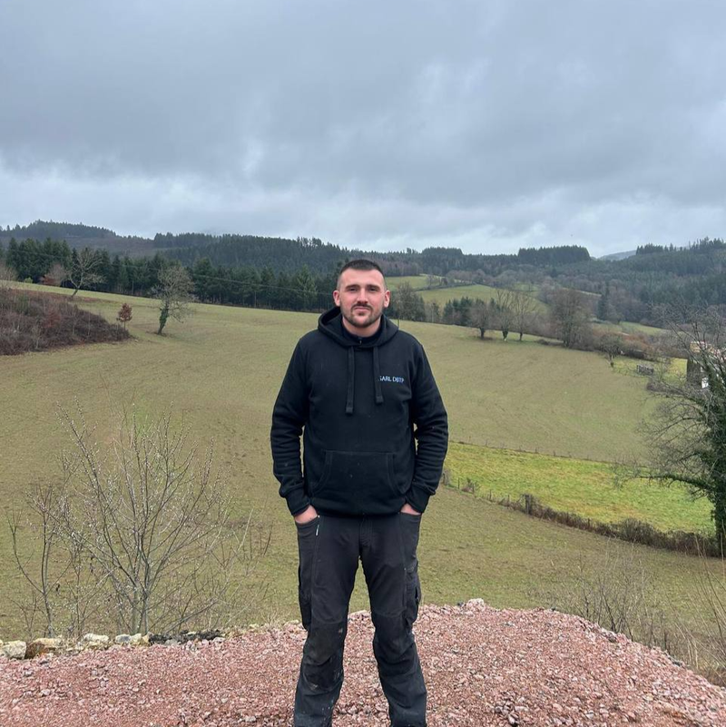 Un homme se tient debout à l'extérieur, sur une colline herbeuse, sous un ciel nuageux, vêtu d'une veste noire et d'un pantalon foncé.