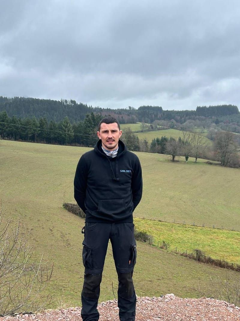 Une personne se tient debout dans un champ vert, entourée de collines boisées, sous un ciel nuageux.