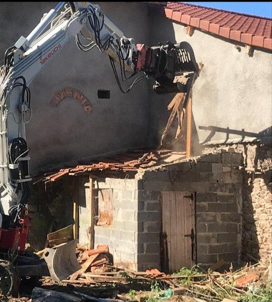 Une excavatrice démolit le mur endommagé d'un bâtiment situé à côté d'un toit de tuiles et d'une petite dépendance