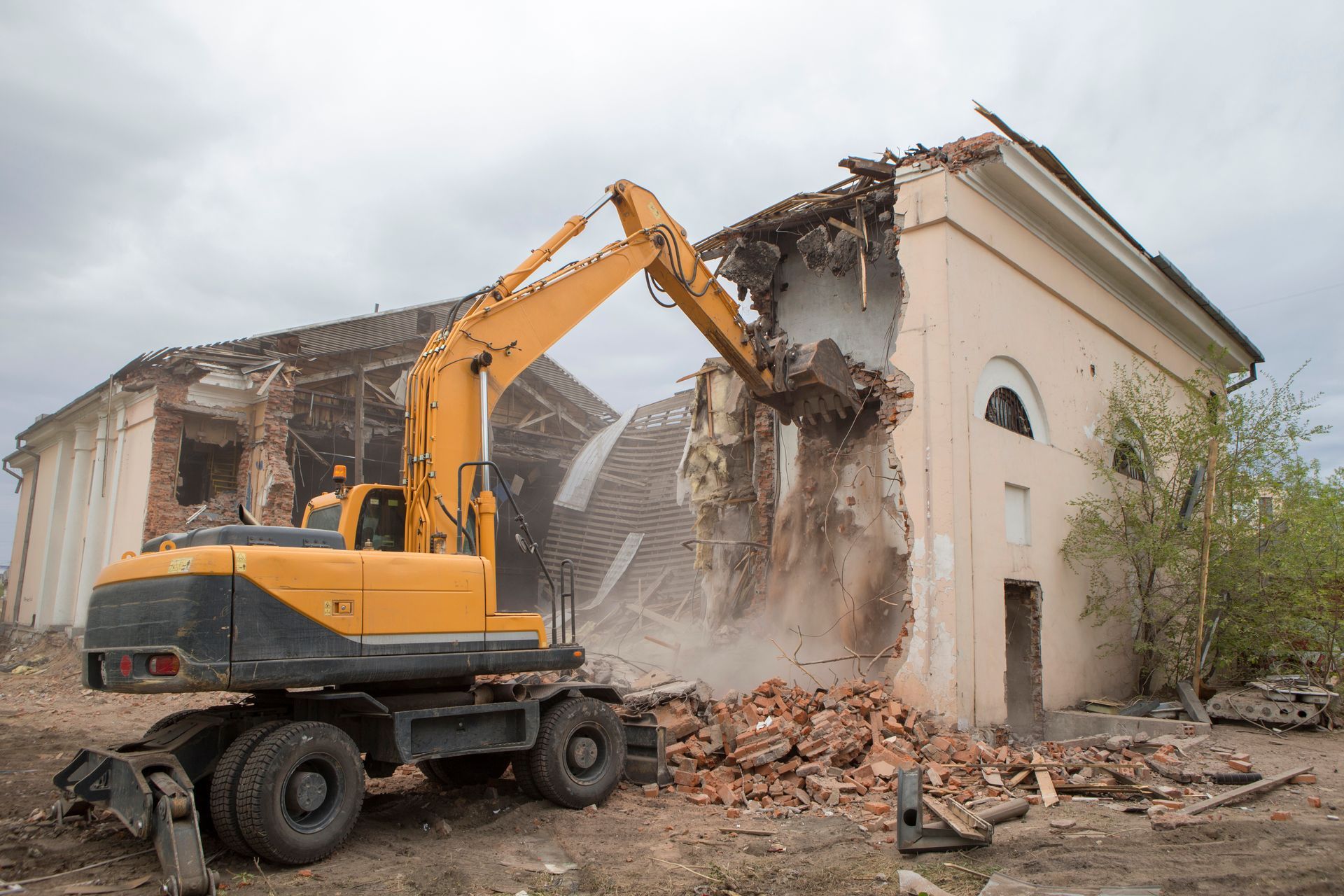 Une pelleteuse jaune démolit un bâtiment beige, des gravats tombent et des débris s'accumulent en dessous.