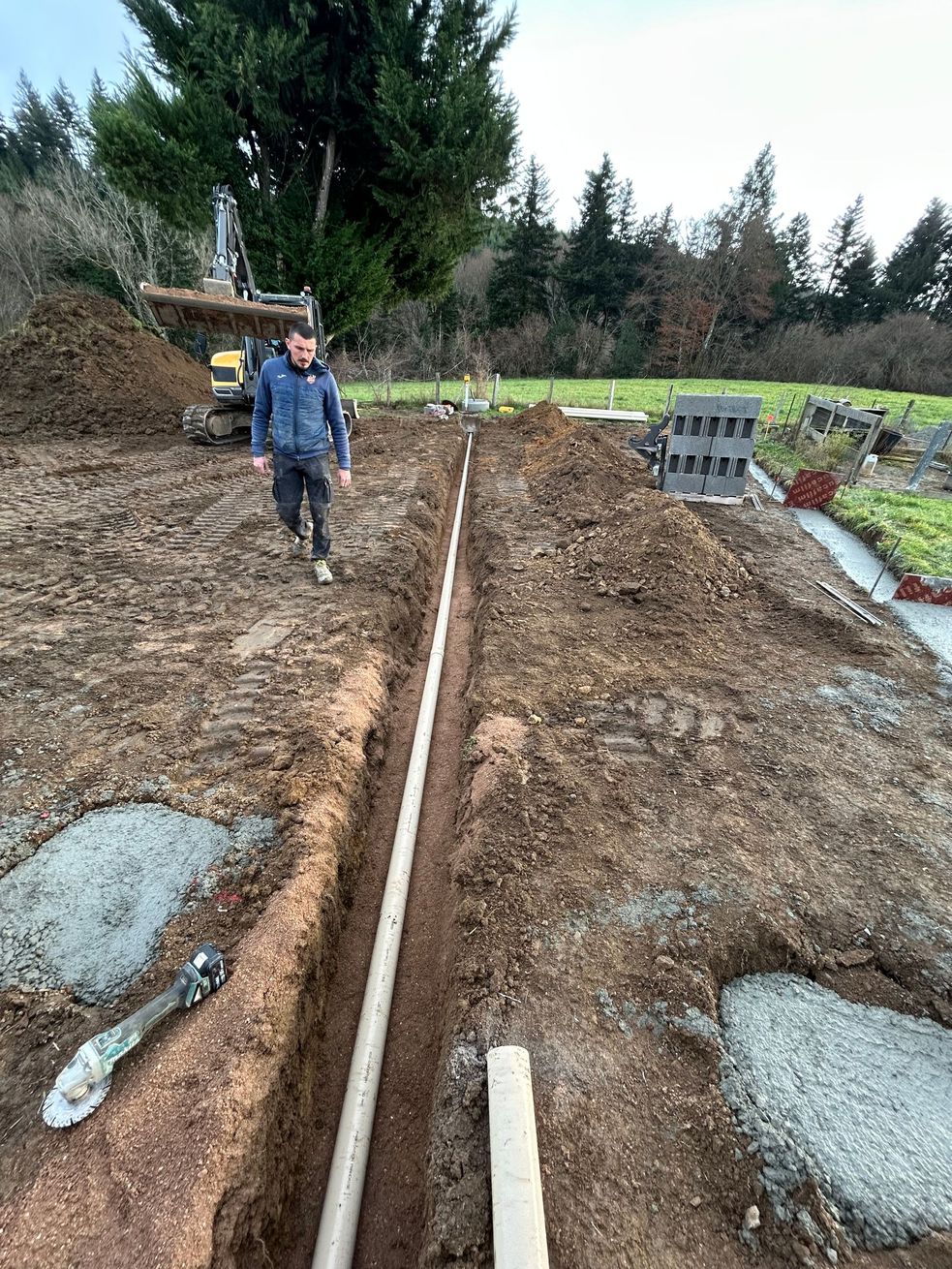 Une tranchée avec un gros tuyau noir est posée à côté d'une excavatrice jaune sur un chantier de construction