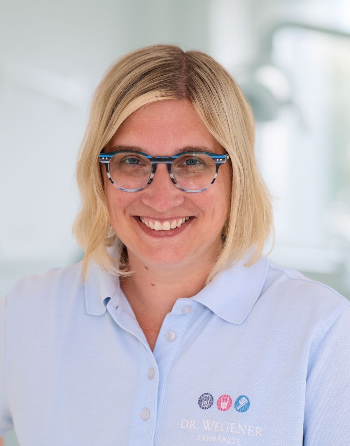 Lächelnde Person mit blonden Haaren und blauer Brille in einem hellblauen Hemd vor einem hellen, verschwommenen Hintergrund