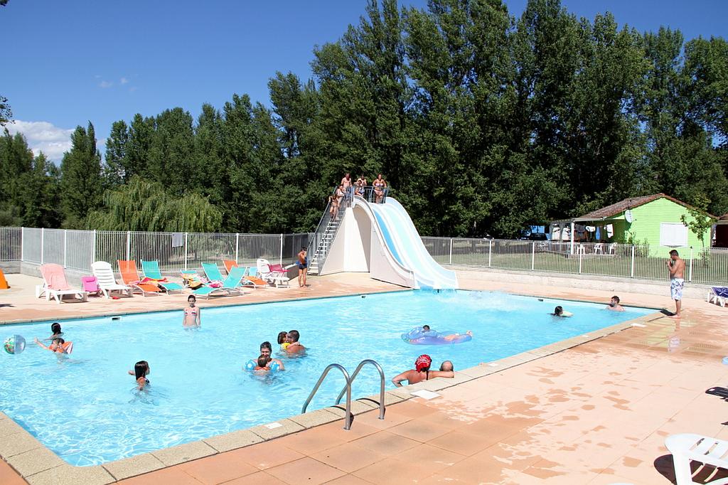 Des personnes nagent et utilisent un toboggan aquatique dans une piscine extérieure ensoleillée, entourée d'arbres et de chaises longues.