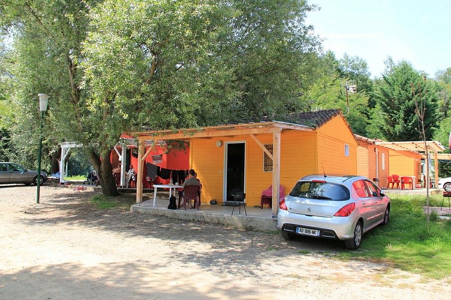 Des cabanes de location orange vif sous de grands arbres, avec une voiture à hayon argentée garée devant, sur un terrain en terre battue.