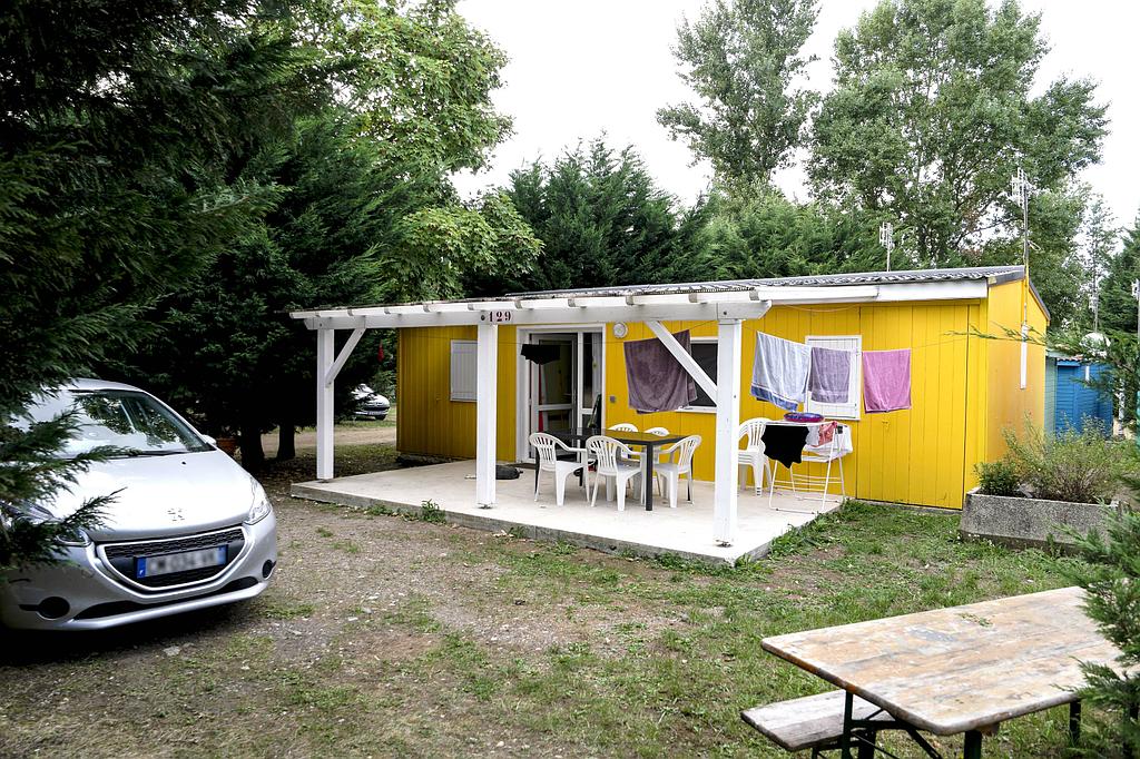 Une cabane jaune vif avec une véranda couverte et du mobilier de jardin, entourée d'arbres, une voiture argentée étant garée à côté.