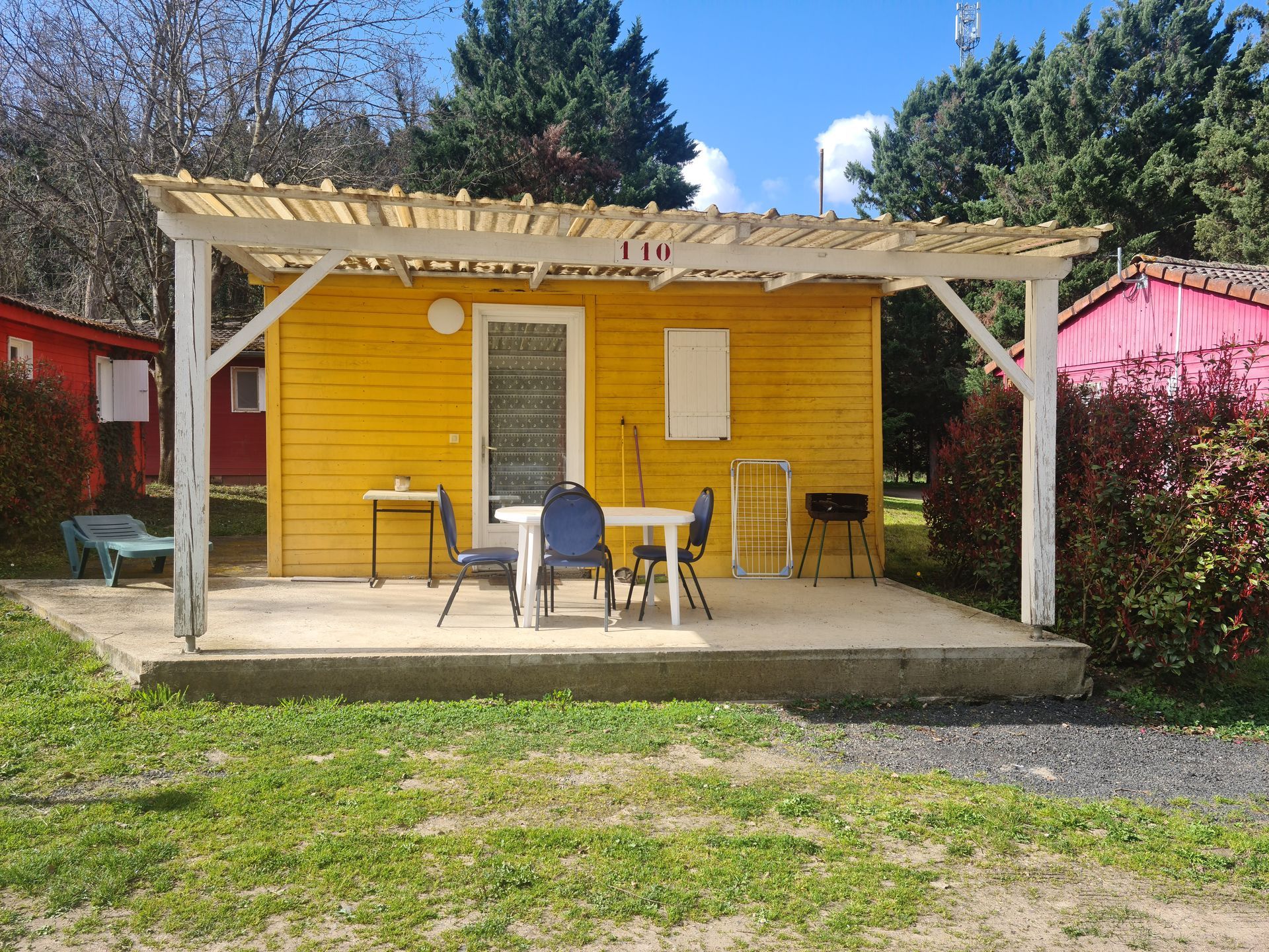 Une cabane en bois jaune avec une terrasse couverte, une petite table, des chaises et un barbecue, sur un terrain de camping herbeux.