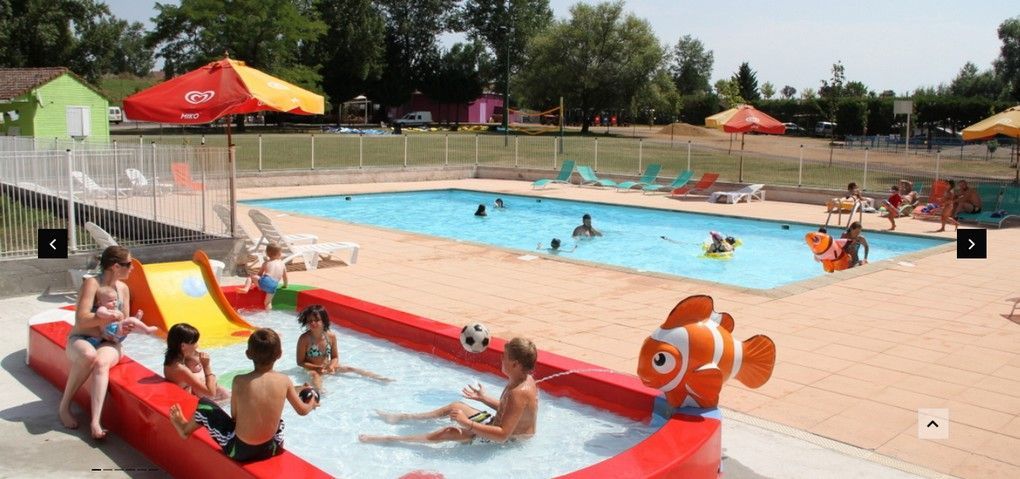 Des enfants jouent dans une pataugeoire rouge peu profonde avec un toboggan et une bouée en forme de poisson, avec une piscine plus grande en arrière-plan.