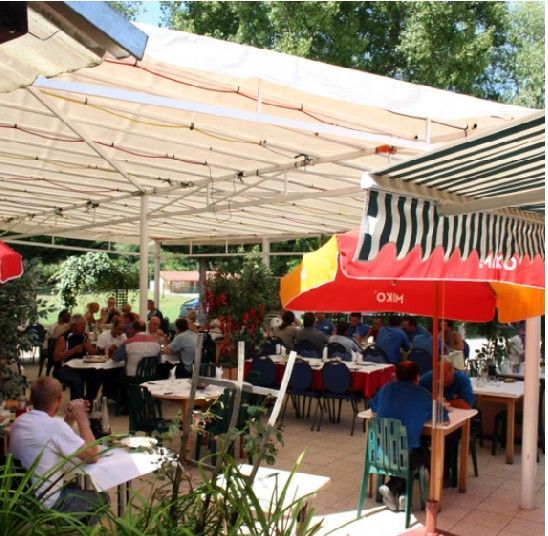 Des personnes dînent à des tables en terrasse, sous des auvents blancs et des parasols rouges, sur un patio ensoleillé bordé d'arbres.
