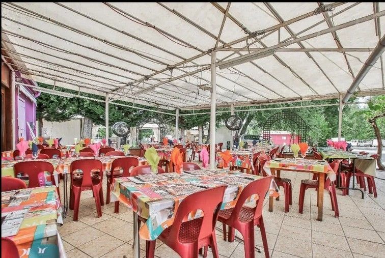 Espace repas extérieur sous un auvent blanc avec des chaises et des tables en plastique rouge recouvertes de nappes à motifs.