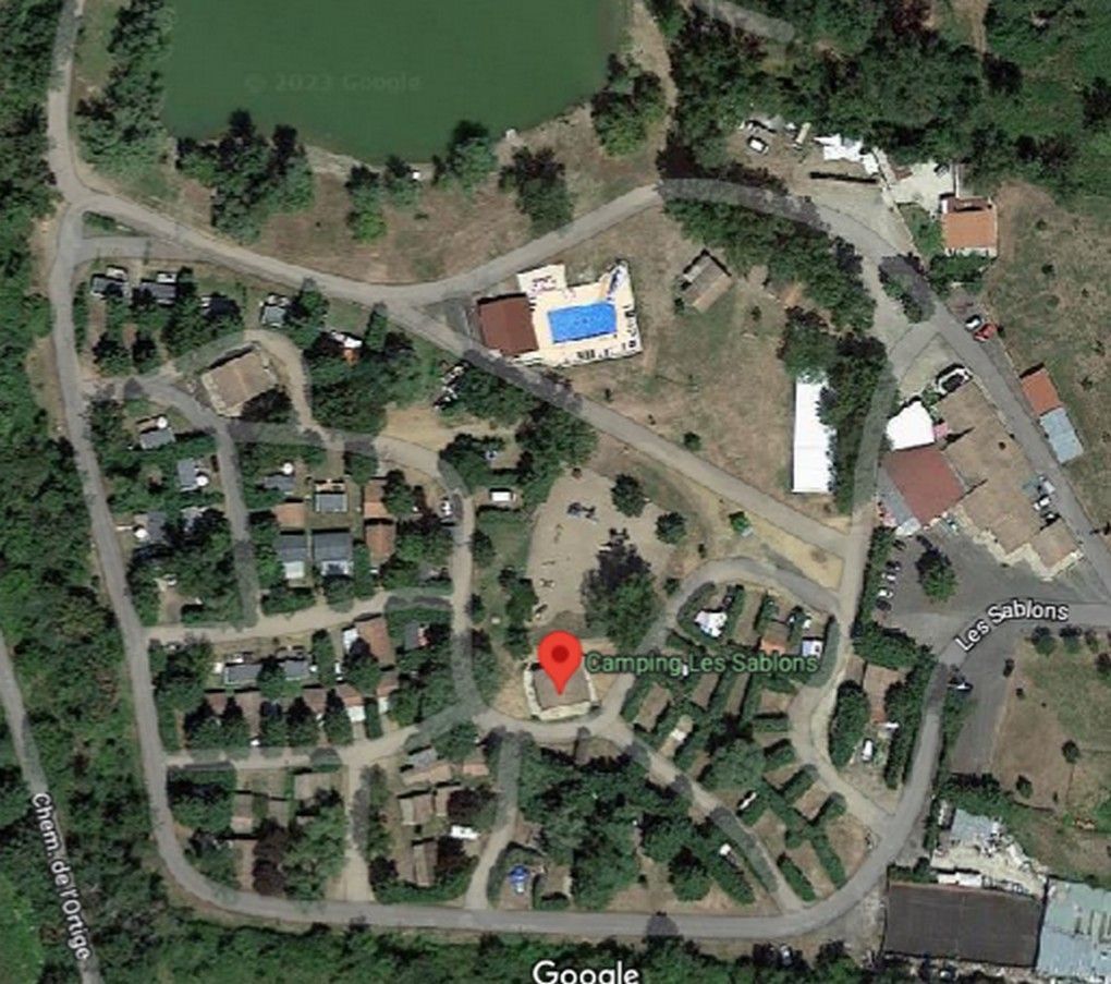 Vue satellite d'un terrain de camping avec de petites cabanes, un bâtiment central marqué par une épingle et une piscine au bord d'un lac.