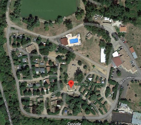 Vue satellite d'un terrain de camping comprenant une piscine, de petits bâtiments de type chalet et un espace central avec une épingle sur la carte.