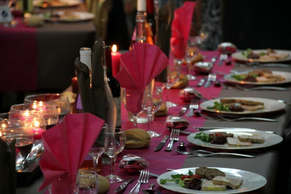 Une table à manger élégante dressée avec des assiettes blanches garnies de mets, des serviettes roses, des bougies et des verres à vin sur une nappe rouge.