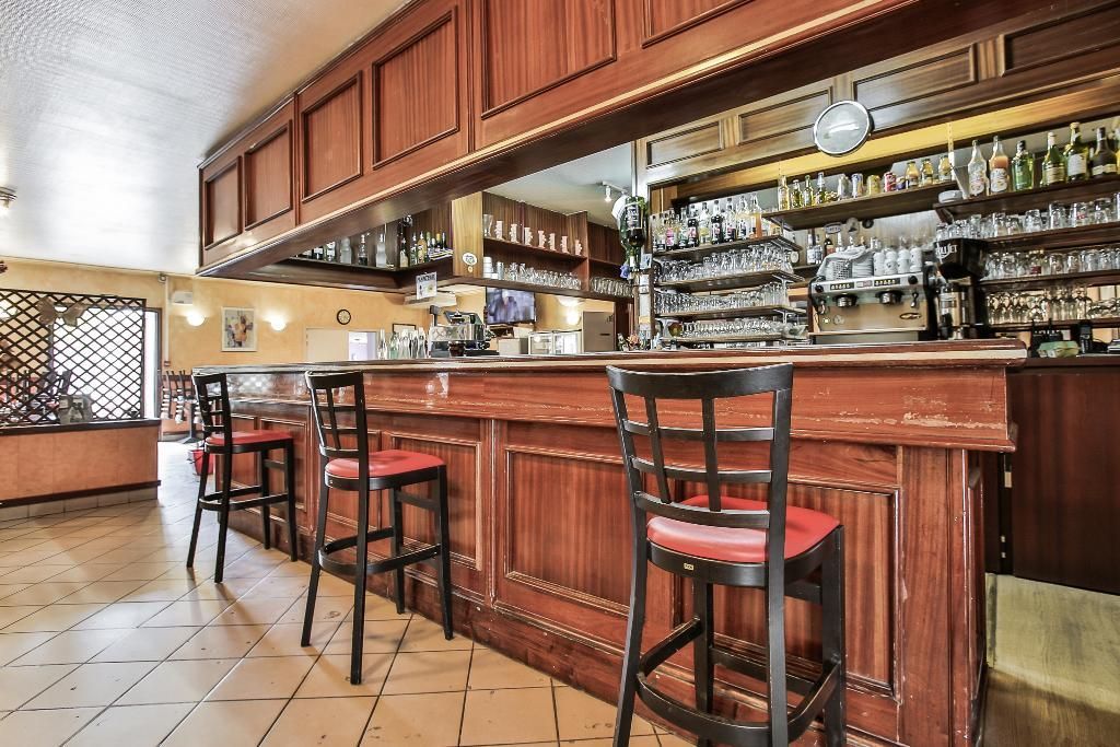 Un intérieur de bar en bois avec trois tabourets hauts, un carrelage à motifs au sol et des étagères garnies de verres et de bouteilles.