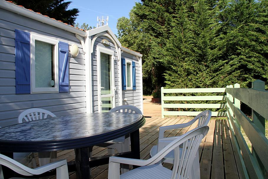 Une caravane bleu clair aux volets bleus est installée sur une terrasse en bois avec une table noire et des chaises en plastique blanc.