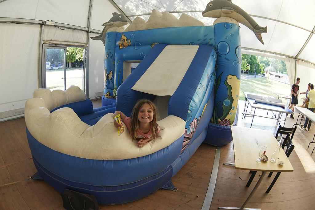 Un enfant sourit à l'intérieur d'un château gonflable bleu sur le thème de l'océan, avec un toboggan, à l'intérieur d'une grande tente.