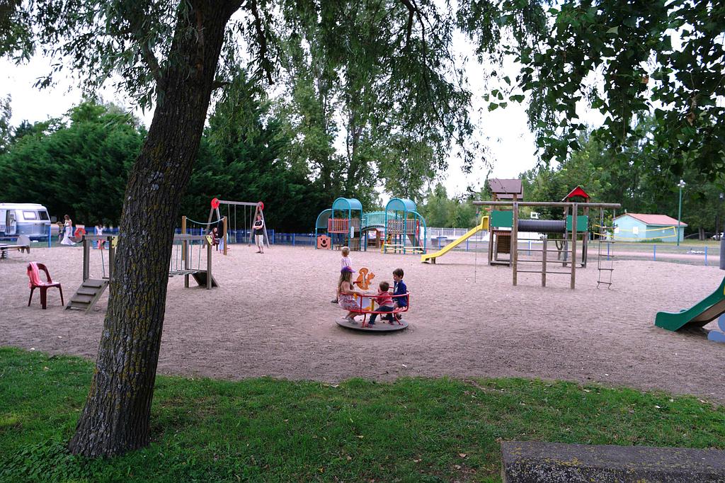 Une aire de jeux dans un parc, avec des enfants qui jouent sur un manège, des balançoires et des structures d'escalade, le tout sur un sol en gravier.