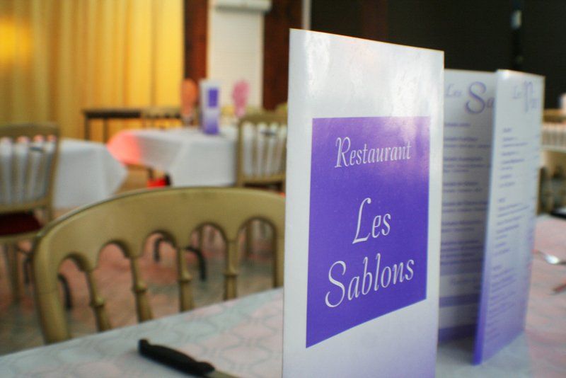 Le menu du restaurant Les Sablons est posé sur une table recouverte d'une nappe dans une salle à manger aux tables nappées de blanc.