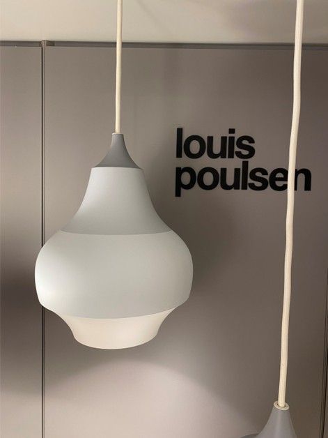 Zwei graue und weiße, tropfenförmige Pendelleuchten hängen vor einer Wand mit einem schwarzen Louis Poulsen-Logo.