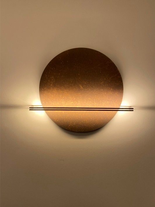 Eine runde, strukturierte Wandleuchte aus Bronze mit einem horizontalen Metallstab erzeugt ein warmes Licht, das an eine schlichte weiße Wand projiziert wird.