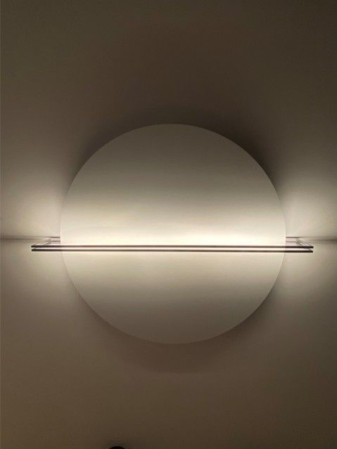 Eine runde, weiße Wandleuchte mit einem horizontalen Metallstab in der Mitte wirft ein sanftes, diffuses Licht an die Wand.