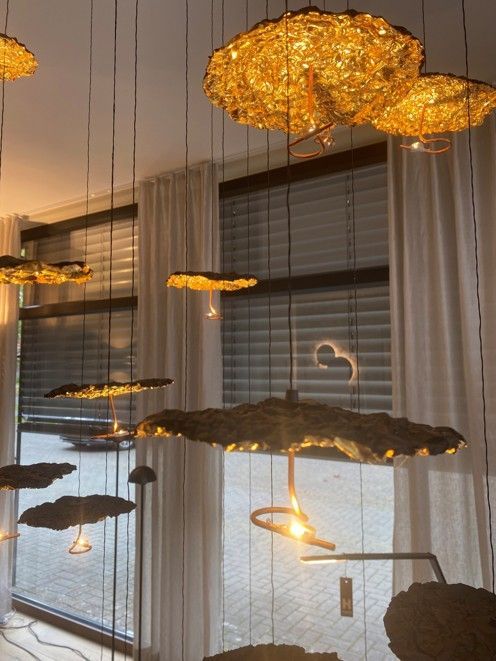 Goldene, strukturierte Pendelleuchten in Form organischer Scheiben hängen in unterschiedlichen Höhen vor einem Fenster mit transparenten Vorhängen.