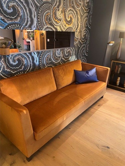 Ein orangefarbenes Samtsofa steht an einer Wand mit gemusterter Tapete und einem Spiegel; auf dem Sitzkissen liegt ein blaues Kissen.