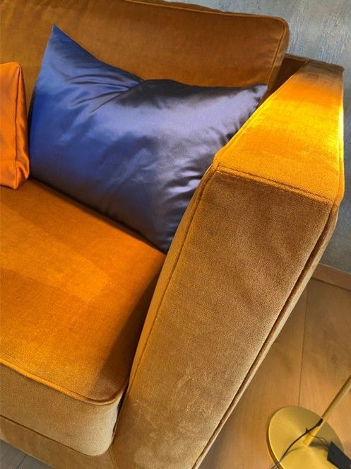 Ein goldfarbenes Samtsofa verfügt über ein großes, glattes, dunkelblaues Zierkissen, das an der Armlehne befestigt ist.