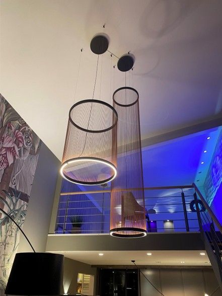 Zwei zylindrische Pendelleuchten im Gitterdesign hängen von der Decke über einem Raum mit Zwischengeschoss.