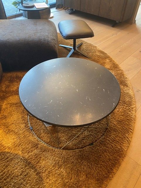 In einem modernen Wohnzimmer steht ein dunkler, runder Couchtisch mit gesprenkelter Tischplatte auf einem runden, zotteligen, senfgelben Teppich.