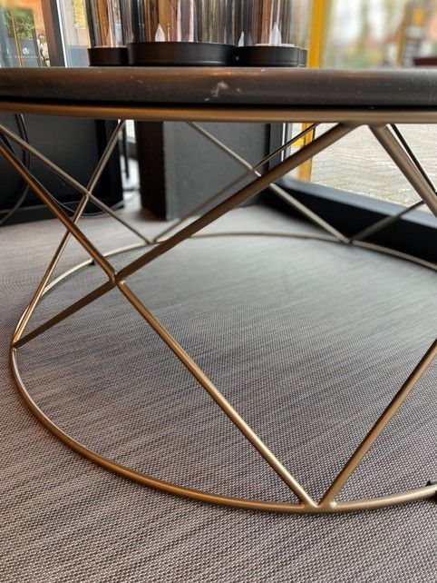 Ein Couchtisch mit dunkler, runder Tischplatte und geometrischem, goldfarbenem Metallgestell steht auf einem grauen Teppichboden.
