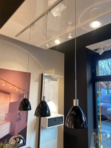 Drei schwarze Pendelleuchten hängen an einer Metallschiene in einem hell erleuchteten Ausstellungsraum mit glänzender Decke und einer Küchenausstellung.