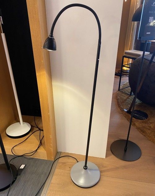 Eine hohe, schlanke schwarze Stehlampe mit flexiblem Schwanenhals und rundem silbernen Fuß steht in einem Ausstellungsraum eines Geschäfts.