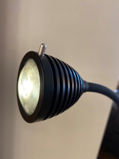 Ein schwarzer, gerippter Scheinwerferkopf an einem flexiblen Arm mit einem kleinen silbernen Schalter obenauf, der weißes Licht ausstrahlt.